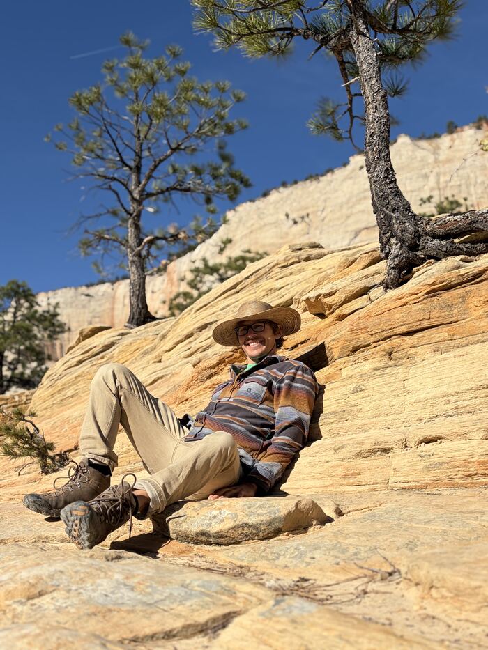 Greg Ellis in a straw hat, smiling and leaning back on sandstone slickrock under a pine tree