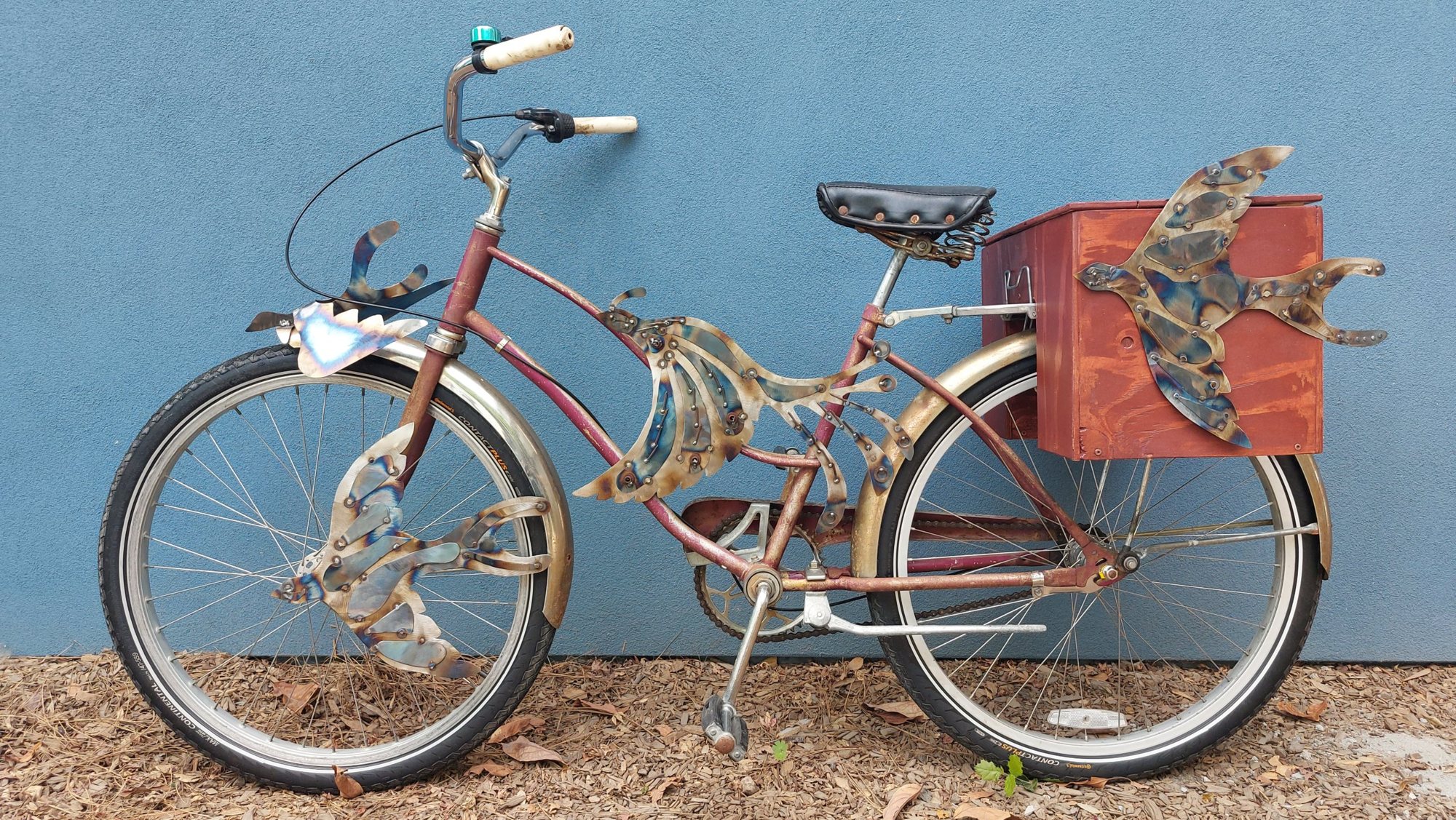 A vintage bicycle against a blue wall, covered in heat-tinted cut-metal birds, with a red wooden art box on the back rack